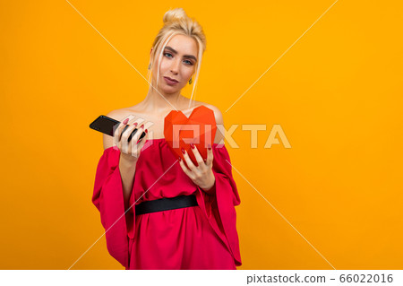 European blond girl in a red dress holds a phone and a red heart made of paper on an orange studio background with copy space 66022016