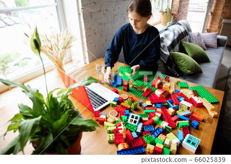Girl playing with constructor at home, watching teacher's tutorial on laptop. Digitalization, remote education Girl playing with constructor at home, watching teacher's tutorial on laptop. Digitalization, remote education 66025839