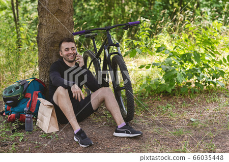 Male cyclist resting under tree after cycling, talking by phone 66035448