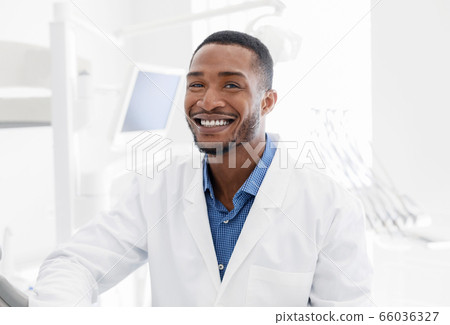 Handsome black dentist smiling over modern dental clinic background Handsome black dentist smiling over modern dental clinic background 66036327