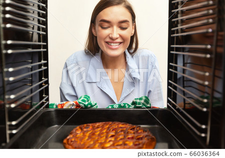 Cheerful millennial housewife taking tasty pie from oven 66036364
