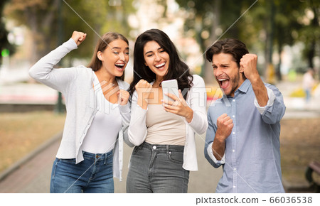 Three Joyful Friends With Cellphone Shaking Fists Celebrating Victory Outdoor 66036538