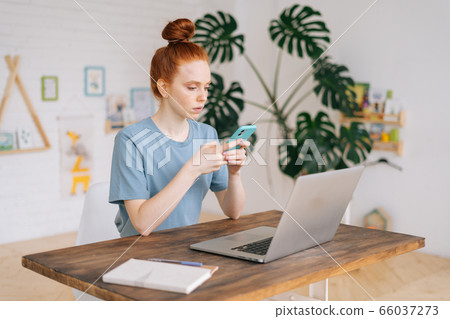 Serious pretty redhead young woman uses the phone while sitting at the table with her laptop. Serious pretty redhead young woman uses the phone while sitting at the table with her laptop. 66037273
