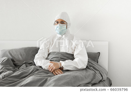Portrait of man in protective white suit and face mask sitting in his bed in his bedroom because of covid-2019 and wait for a end of coronavirus quarantine. Self isolation. 66039796