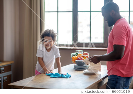 Small cute curly-haired mulatta wiping the table surface and smiling, her father standing 66051019