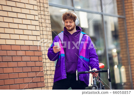 Handsome young man using mobile phone and headphones while standing near his bicycle beside him Handsome young man using mobile phone and headphones while standing near his bicycle beside him 66058857