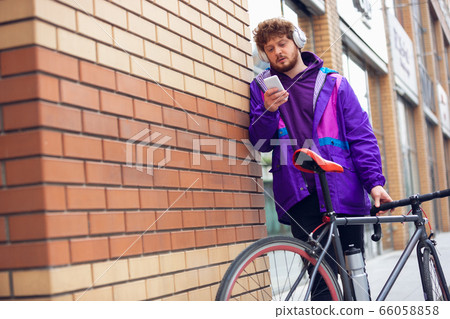 Handsome young man using mobile phone and headphones while standing near his bicycle beside him Handsome young man using mobile phone and headphones while standing near his bicycle beside him 66058858