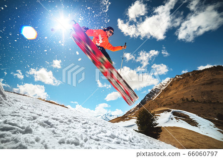 A young stylish man in sunglasses and a cap performs a trick in jumping with a kicker of snow against the blue sky and the sun on a sunny day. The concept of park skiing in winter or spring 66060797
