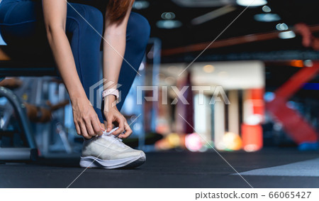 Young Asian Women are tying shoelaces To prepare before exercise and for safety, A healthy woman sitting on the bench leaning down to tie the shoelace at gym with blurback ground and copy space Young Asian Women are tying shoelaces To prepare before exercise and for safety, A healthy woman sitting on the bench leaning down to tie the shoelace at gym with blurback ground and copy space 66065427