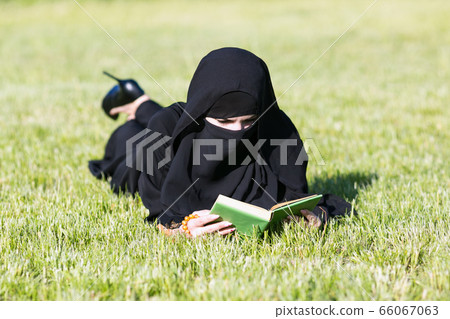 Islamic woman reads a book on a lawn in a park. Islamic woman reads a book on a lawn in a park. 66067063