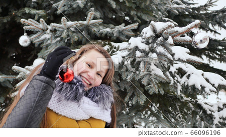 Attractive young girl with long hair in a winter suit posing against a snowy tree. A girl is stringing Christmas balls on a branch. Winter in the forest. Attractive young girl with long hair in a winter suit posing against a snowy tree. A girl is stringing Christmas balls on a branch. Winter in the forest. 66096196