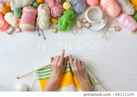 Hands of craftswoman knit a bright knitted striped fabric. Woolen and cotton yarn lies in a basket. Cappuccino coffee, cookies and glasses lie on a white wooden background. 66111124