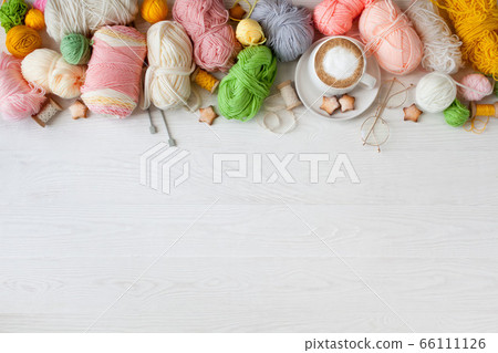 Knitted bright striped fabric. Woolen and cotton yarn lies in a basket. Cappuccino coffee, cookies and glasses lie on a white wooden background. 66111126