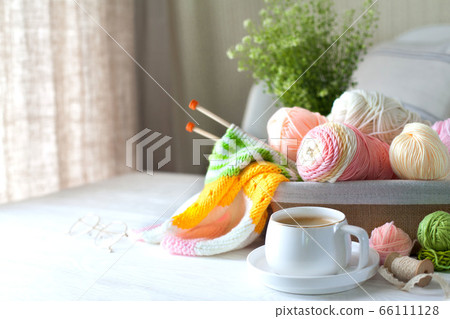 Knitted bright striped fabric. Woolen and cotton yarn lies in a basket. Cappuccino coffee, cookies and glasses lie on a white wooden background. 66111128