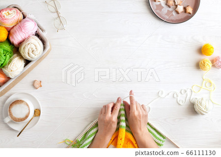 Hands of craftswoman knit a bright knitted striped fabric. Woolen and cotton yarn lies in a basket. Cappuccino coffee, cookies and glasses lie on a white wooden background. 66111130