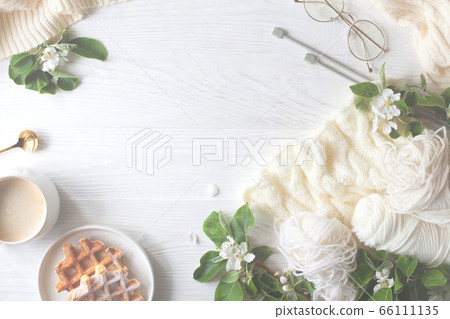 Knitting, knitting needles, yarn, coffee and waffles stand on a white wooden background. Surrounded by branches of a blossoming apple tree. Flatlay. 66111135