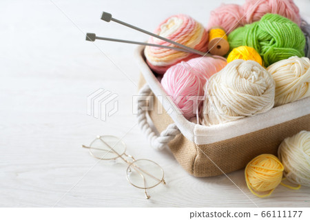 Woolen and cotton bright yarn lies in a basket. Glasses lie on a white wooden background. Woolen and cotton bright yarn lies in a basket. Glasses lie on a white wooden background. 66111177