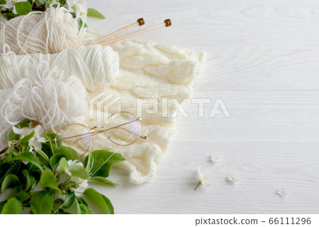 Knitting, knitting needles, yarn and glasses lie on a white wooden background. Surrounded by branches of a blossoming apple tree. 66111296