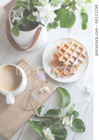 A bouquet of branches of a blossoming apple tree stands in a white vase on the table. Viennese waffles on a plate. Coffee in a white cup. Flatlay 66111298