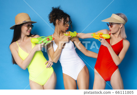 Three cheerful girlfriends in bikinis playing with water guns over blue background Three cheerful girlfriends in bikinis playing with water guns over blue background 66111805