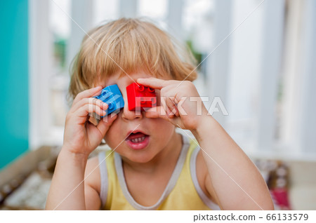 The boy makes eyes of colorful children's blocks. Cute little kid boy with glasses playing with lots of colorful plastic blocks indoor. Promotion of skills and creativity 66133579