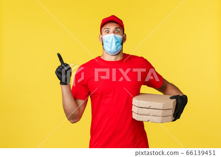 Food delivery, tracking orders, covid-19 and self-quarantine concept. Shocked young courier in red uniform and medical mask, pointing finger upper left corner as deliver pizza to clients house 66139451