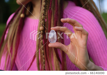 Adult girl in knitted sweater with long dreadlocks is holding transparent violet amethyst yoni egg for vumfit, imbuilding or meditation outdoors on her body background outdoors 66140890