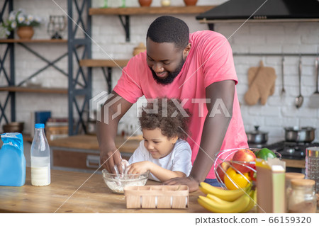 African american bearded man playing with his daughter stirring the flour in the bowl 66159320