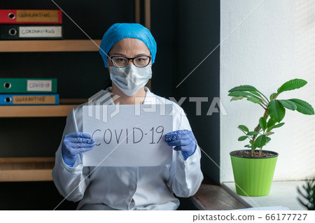 Doctor with inscription covid-19 in hands. Serious Asian woman in protective uniform looking at the camera while sitting in medical office interior. Quarantine concept 66177727