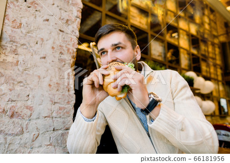 Side angle view of young handsome hungry man eating croissant while sitting in outdoor cafe. Portrait of handsome young Caucasian man eating delicious croissant 66181956