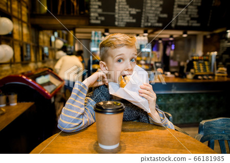 Happy cute boy eating fresh croissant, sitting at the table in city cafe, with paper cup of tea or cocoa Happy cute boy eating fresh croissant, sitting at the table in city cafe, with paper cup of tea or cocoa 66181985