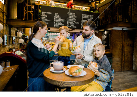 Young cheerful family, bearded father, pretty mother and two children boys, drinking hot chocolate drink and eating traditional French croissants in a cozy indoor city cafe. Young cheerful family, bearded father, pretty mother and two children boys, drinking hot chocolate drink and eating traditional French croissants in a cozy indoor city cafe. 66182012