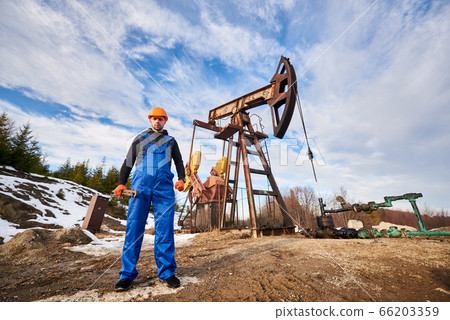 Oil worker in uniform and helmet working in an oilfield next to a pump jack on a sunny day Oil worker in uniform and helmet working in an oilfield next to a pump jack on a sunny day 66203359