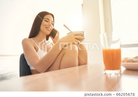 Smiling young woman having light breakfast and speaking on phone Smiling young woman having light breakfast and speaking on phone 66205678