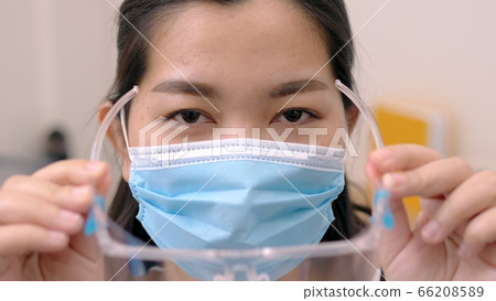 Portrait of an Asia young female who is wearing a face shield with mask rounded around her face from a frontal perspective to protect her glasses and eyes prepare to new normal. Coronavirus pandemic. Portrait of an Asia young female who is wearing a face shield with mask rounded around her face from a frontal perspective to protect her glasses and eyes prepare to new normal. Coronavirus pandemic. 66208589