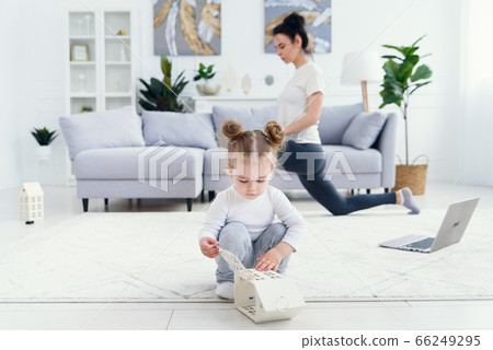 Funny baby girl playing at home while her sporty healthy mommy doing fitness and yoga exercises on the background. Funny baby girl playing at home while her sporty healthy mommy doing fitness and yoga exercises on the background. 66249295