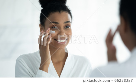 Mirror reflection smiling African American woman applying moisturizing cream Mirror reflection smiling African American woman applying moisturizing cream 66258387