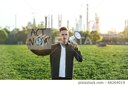 Young activist with placard and megaphone standing outdoors by oil refinery, protesting. 66264019