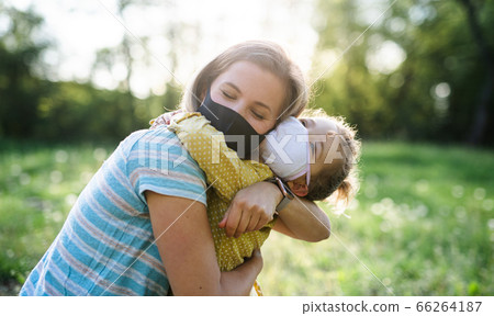 Side view of happy mother hugging small daughter in nature, wearing face masks. Side view of happy mother hugging small daughter in nature, wearing face masks. 66264187