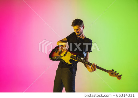 Young caucasian musician playing bass guitar in neon light on pink-green background Young caucasian musician playing bass guitar in neon light on pink-green background 66314331