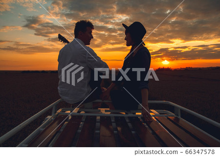 Silhouette of a man with a guitar and a woman in a hat sitting on the roof of a car on the background of the sunset. Silhouette romance concept. 66347578