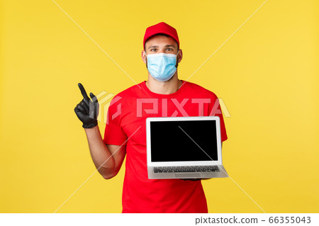 covid-19, safe shopping, express delivery and order tracking concept. Excited young courier in medical mask and red uniform, pointing finger left at promo, showing laptop screen, link or webpage 66355043