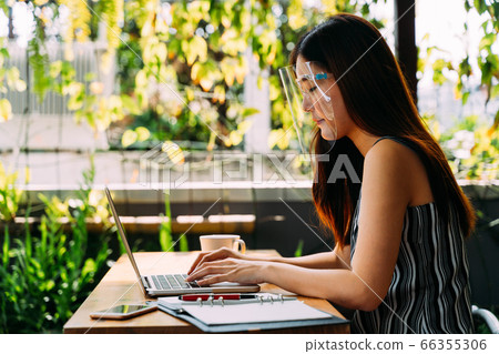 Asian woman wearing a face shield outside while sitting and working on laptop. Using computer in outdoor office. Face shield is popular equipment to prevent Corona Virus in Asia Asian woman wearing a face shield outside while sitting and working on laptop. Using computer in outdoor office. Face shield is popular equipment to prevent Corona Virus in Asia 66355306