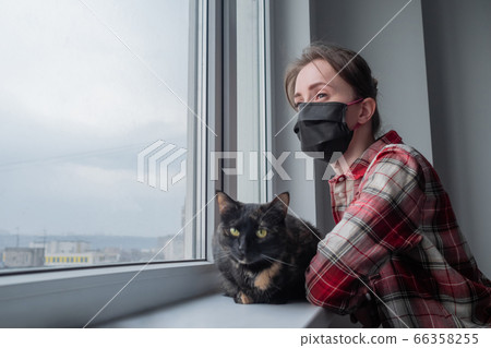 Woman in medical face mask and black cat looking out of window - side view Woman in medical face mask and black cat looking out of window - side view 66358255