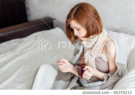 A sick young woman, sitting in bed with a scarf, suffers from a sore throat. Drinking hot tea. A woman with a fever wrapped in a blanket. A sick young woman, sitting in bed with a scarf, suffers from a sore throat. Drinking hot tea. A woman with a fever wrapped in a blanket. 66359213