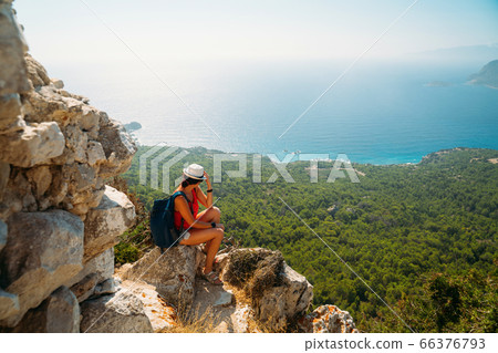 Happy woman traveler is siting next to Monolithos castle and admires the picturesque view of the Mediterranean Sea 66376793