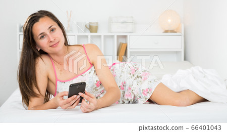 Smiling girl resting and using smartphone on bed in bedroom Smiling girl resting and using smartphone on bed in bedroom 66401043
