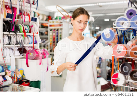 Interested young woman choosing colorful ribbons and braid for dressmaking in sewing supplies shop 66401151