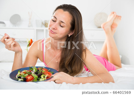 Positive pretty woman holding fork and eating vegetable salad in bed 66401569
