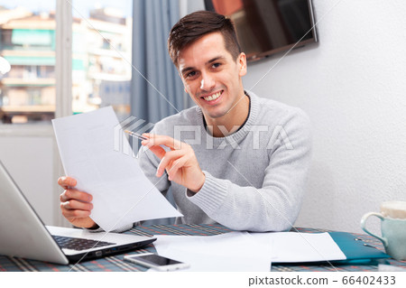 Young happy man sitting at home table working with papers, analyzing domestic budget Young happy man sitting at home table working with papers, analyzing domestic budget 66402433